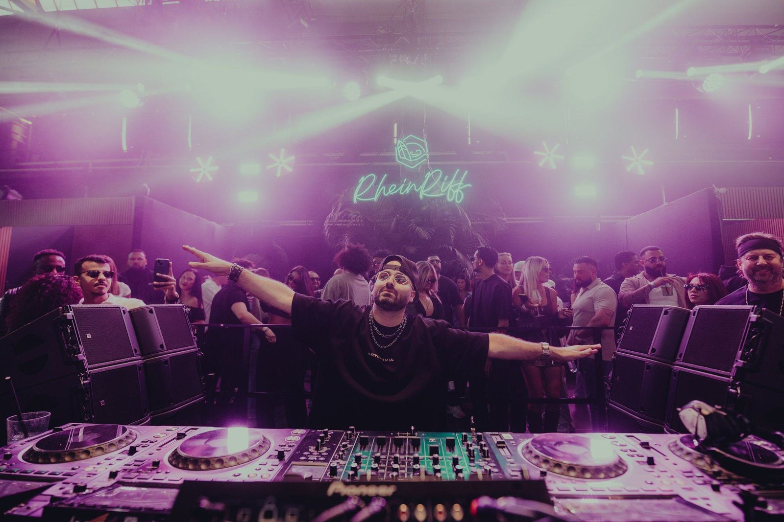 ERAN with his arms spread wide behind the decks in a packed club under purple lights